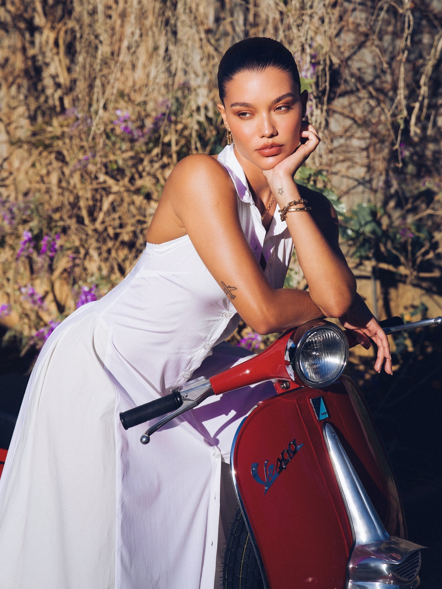 Agnes de vestido branco longo casual apoiada em uma moto Vespa vermelha clássica